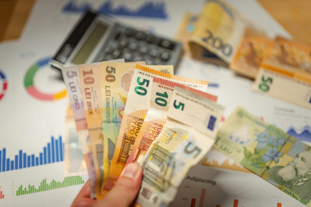 Een hand houdt verschillende coupures van Europese bankbiljetten vast boven financiële grafieken. Een rekenmachine is zichtbaar op de achtergrond, gedeeltelijk bedekt door meer bankbiljetten. De grafieken tonen kleurrijke grafieken en data.