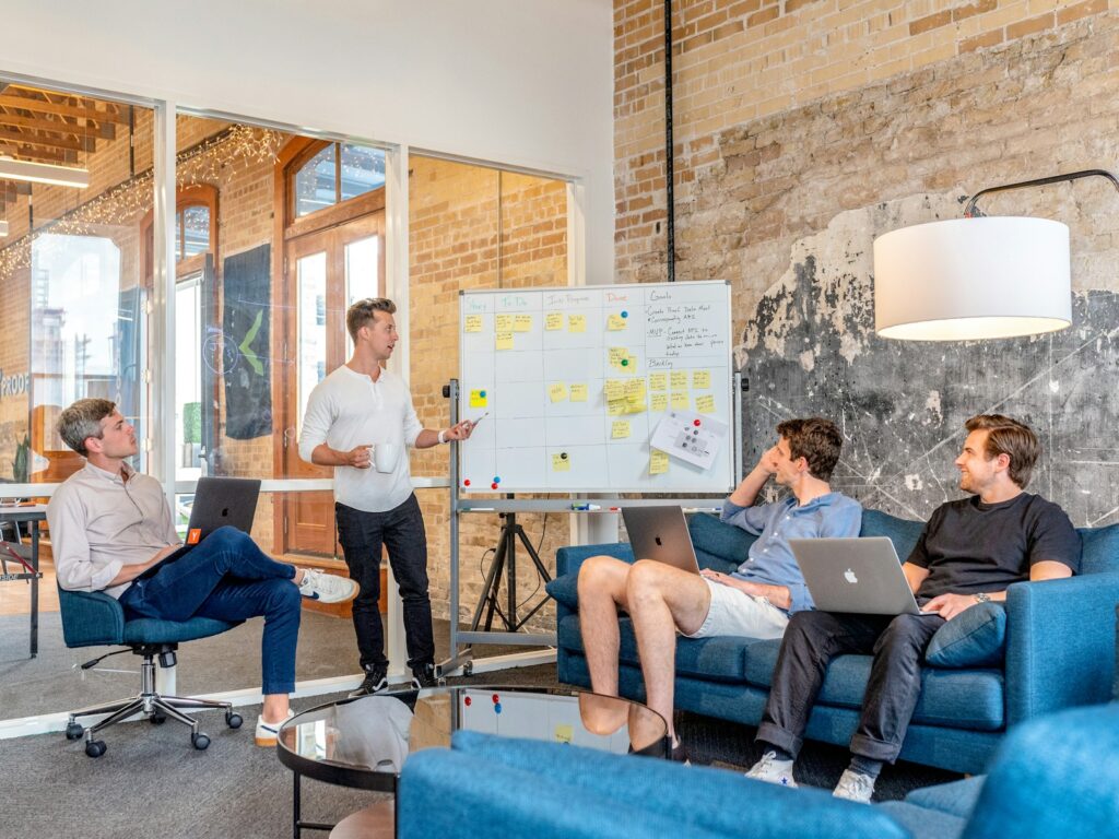 Vier mensen zitten in een kamer met bakstenen muren. Drie zitten op blauwe banken met laptops, terwijl één persoon staat en naar een whiteboard wijst dat vol zit met plaknotities. Een grote staande lamp staat in de hoek en op de achtergrond is een glazen wand.