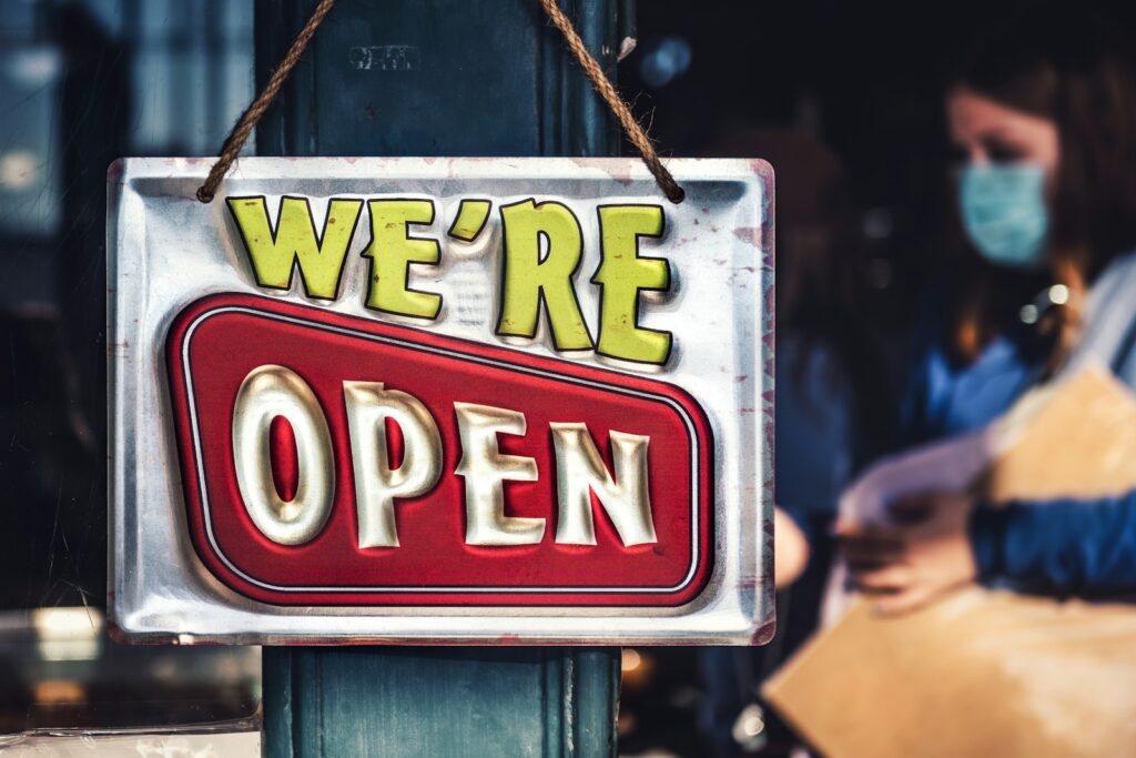 Een metalen bord met de woorden "WE'RE OPEN" hangt buiten een winkelpui. Het bord heeft een retro-ontwerp met felgele en rode letters.