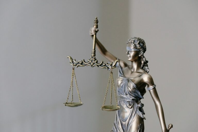 Een bronzen beeld van Lady Justice met een weegschaal in haar rechterhand. Ze is geblinddoekt en draagt een vloeiende jurk. De achtergrond is een effen, lichtgekleurde muur.