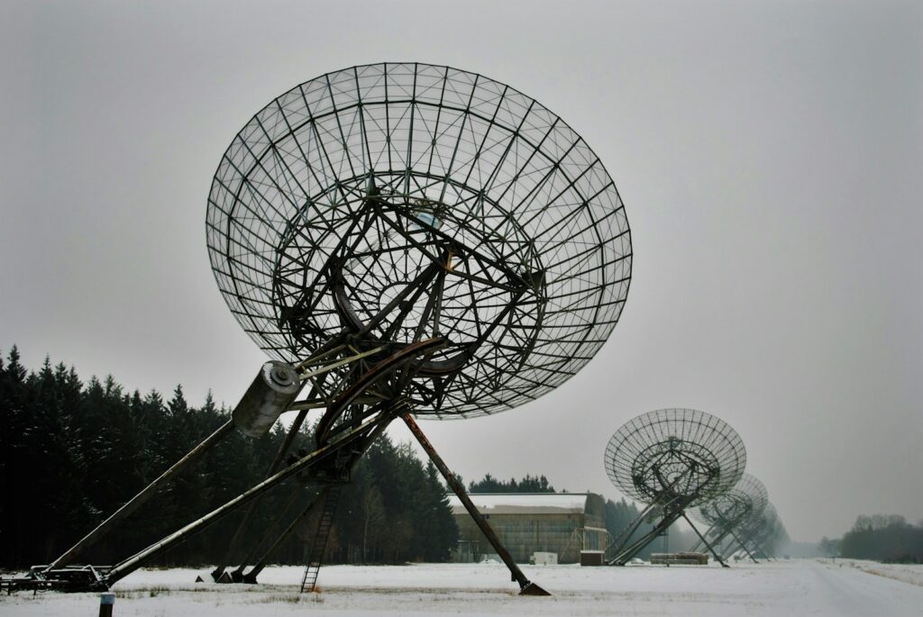 Een rij grote radiotelescopen staat in een besneeuwd veld, gericht op de lucht. De schotels zijn omringd door een bos en een gebouw is zichtbaar op de achtergrond onder een bewolkte lucht.