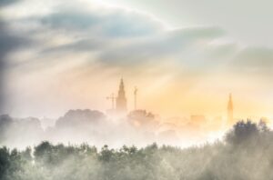 city, groningen, dutch, holland, De skyline van een stad met hoge gebouwen en kranen wordt gedeeltelijk bedekt door ochtendmist en bomen, terwijl op de achtergrond het zonlicht door de wolken schijnt.