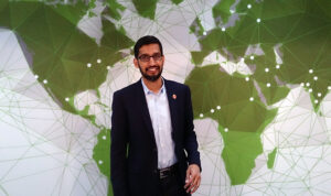 Een man in een donker pak en een bril staat voor een muur met een groene wereldkaart met verbindende lijnen en stippen. Sundar Pichai CEO Google Deepmind