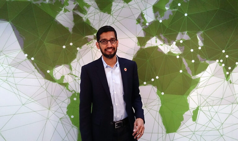 Een man in een donker pak en een bril staat voor een muur met een groene wereldkaart met verbindende lijnen en stippen. Sundar Pichai CEO Google Deepmind