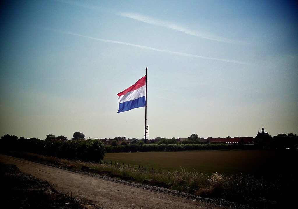 Een grote rood-wit-blauw gestreepte vlag hangt halfstok aan een hoge vlaggenmast in een landelijk gebied, met bomen, struiken en een paar gebouwen op de achtergrond, onder een heldere hemel.