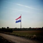 Een grote rood-wit-blauw gestreepte vlag hangt halfstok aan een hoge vlaggenmast in een landelijk gebied, met bomen, struiken en een paar gebouwen op de achtergrond, onder een heldere hemel.