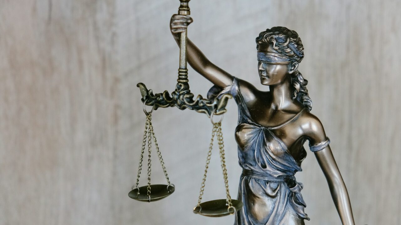 Een bronzen beeld van Vrouwe Justitia met in haar linkerhand een weegschaal en in haar rechterhand een zwaard. Ze draagt een blinddoek en staat tegen een lichte, wazige achtergrond.