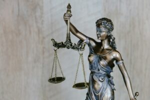 Een bronzen beeld van Vrouwe Justitia met in haar linkerhand een weegschaal en in haar rechterhand een zwaard. Ze draagt een blinddoek en staat tegen een lichte, wazige achtergrond.