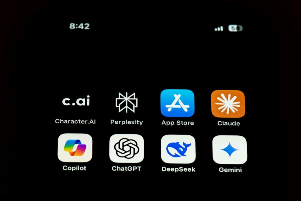 Op een smartphonescherm worden acht app-iconen weergegeven: Character.AI, Perplexity, App Store, Claude, Copilot, ChatGPT, DeepSeek en Gemini. De tijd linksboven is 8:42 en de batterij- en signaaliconen rechtsboven.
