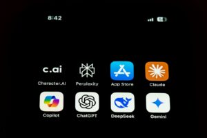 Op een smartphonescherm worden acht app-iconen weergegeven: Character.AI, Perplexity, App Store, Claude, Copilot, ChatGPT, DeepSeek en Gemini. De tijd linksboven is 8:42 en de batterij- en signaaliconen rechtsboven.