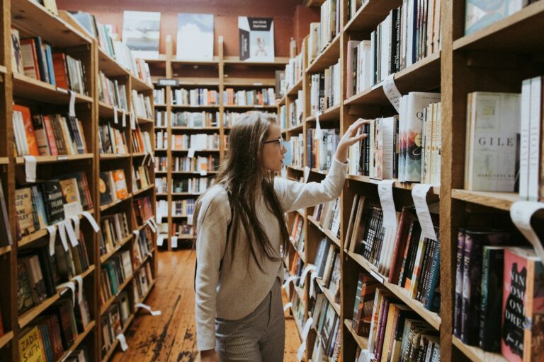 Een vrouw met lang bruin haar en een bril staat in een boekwinkel en reikt naar een boek van een plank. De houten planken aan beide kanten staan vol met boeken en er hangen handgeschreven briefjes aan de planken.