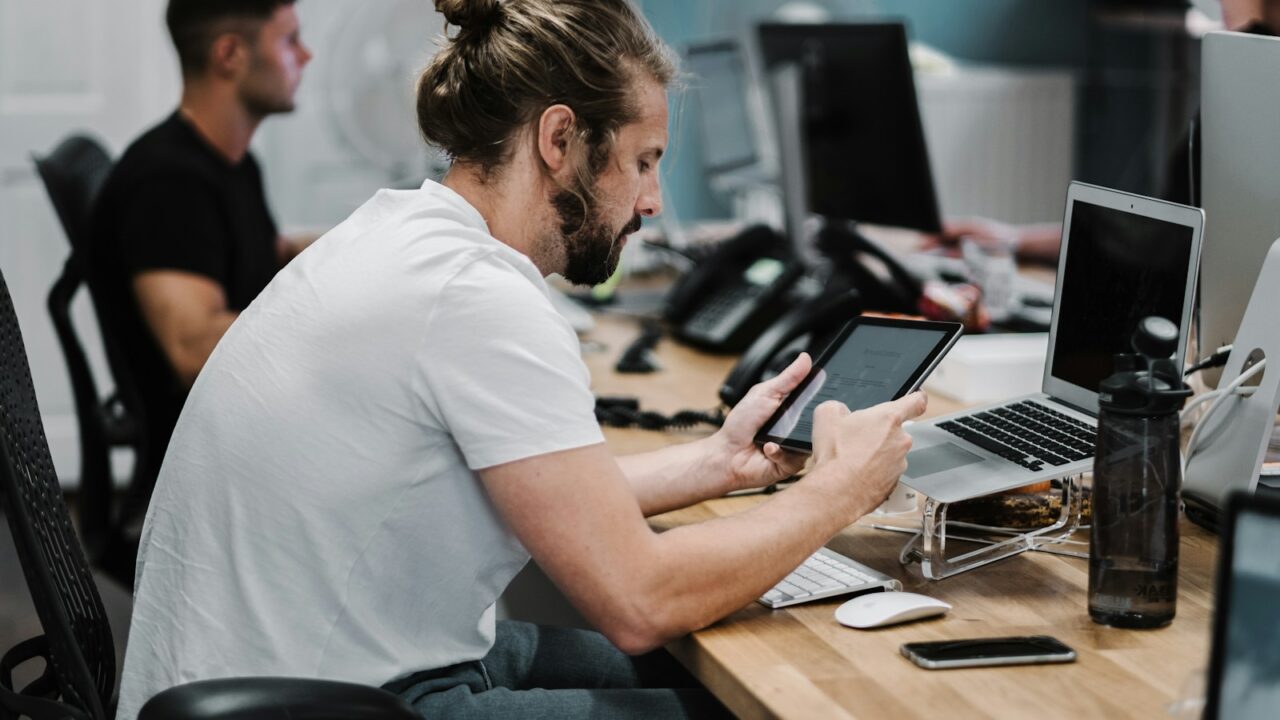 Een man met een baard en een samengebonden haar zit achter een bureau met een tablet, met een opengeklapte laptop en diverse kantoorbenodigdheden in de buurt. Een andere persoon werkt aan een nabijgelegen bureau op de achtergrond.