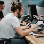 Een man met een baard en een samengebonden haar zit achter een bureau met een tablet, met een opengeklapte laptop en diverse kantoorbenodigdheden in de buurt. Een andere persoon werkt aan een nabijgelegen bureau op de achtergrond.