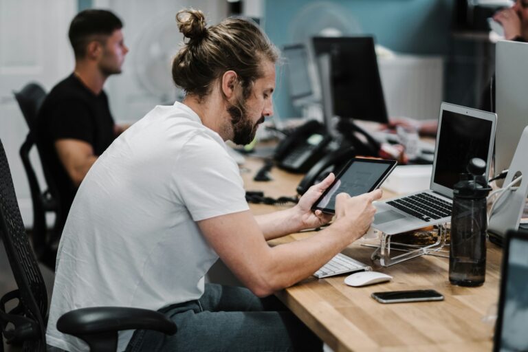 Een man met een baard en een samengebonden haar zit achter een bureau met een tablet, met een opengeklapte laptop en diverse kantoorbenodigdheden in de buurt. Een andere persoon werkt aan een nabijgelegen bureau op de achtergrond.