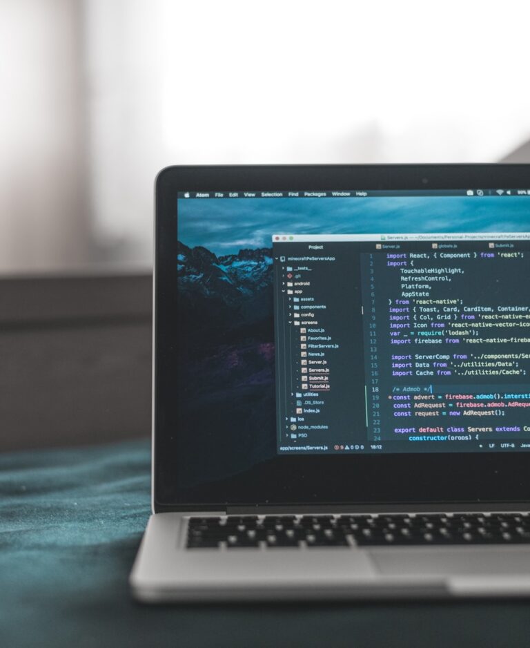Een laptop met een code-editor, JavaScript-bestanden en -mappen geopend en coderegels op het scherm. De achtergrond is wazig en de laptop staat op een donkere ondergrond.